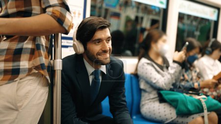 Foto de Sonriente guapo inversor masculino escuchando música relajante mientras está sentado en el tren. El hombre de negocios profesional disfruta escuchando música mientras usa el transporte público para ir al lugar de trabajo. Fondo borroso. Exultante. - Imagen libre de derechos
