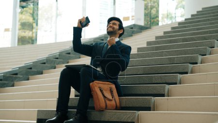 Photo for Successful business man celebrate successful project while sitting at stairs. Smart project manager getting new gob, getting promotion, increasing sales while calling friends by using phone. Exultant. - Royalty Free Image