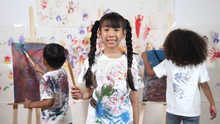 Photo for Creative cute girl pose at camera while diverse children painting behind. Young beautiful child looking at camera while standing at stained room and holding paintbrush. Creative activity. Erudition. - Royalty Free Image