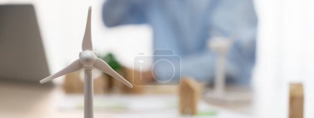 Photo for Windmill model represented using clean energy and wooden block represented eco house was scatter around on the table in front of businessman working on laptop. Blurring background. Delineation. - Royalty Free Image