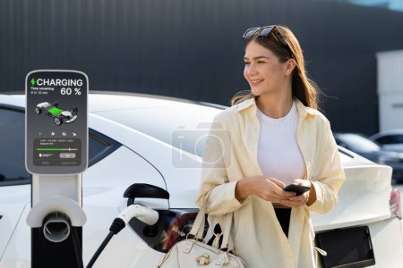 Photo for Young woman holding shopping bag and use smartphone to pay for electricity for recharging EV car battery from charging station at city mall parking lot. Modern woman go shopping by eco car. Expedient - Royalty Free Image