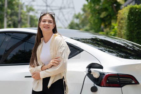 Téléchargez les photos : Jeune femme recharger son véhicule électrique EV au parking vert du parc de la ville. Style de vie urbain durable pour voiture écologique EV avec station de charge de la batterie. Expedient - en image libre de droit