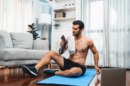 Photo for Athletic and sporty man drinking water on fitness mat after finishing home body workout exercise session for fit physique and healthy sport lifestyle at home. Gaiety home exercise workout training. - Royalty Free Image