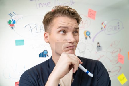 Photo for Portrait of young caucasian businessman thinking with confused face expression while standing in front of glass board with sticky notes and mind map at creative business meeting. Immaculate. - Royalty Free Image