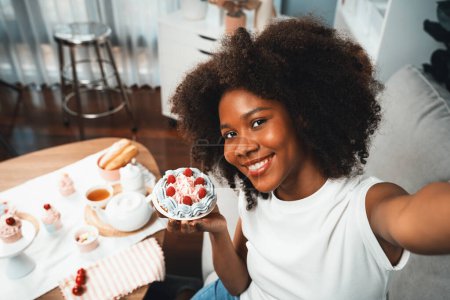 Téléchargez les photos : Belle jeune blogueuse africaine présentant morceau de cupcake en concept cuisine spéciale avec selfie sur smartphone. Création de contenu de médias sociaux avec plat préféré de boulangerie de bonbons. Tastemaker. - en image libre de droit