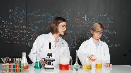 Photo for Caucasian teacher helps student mixed liquid in science laboratory. Professional instructor support smart boy doing experiment in STEM science class at blackboard written chemical theory. Erudition. - Royalty Free Image