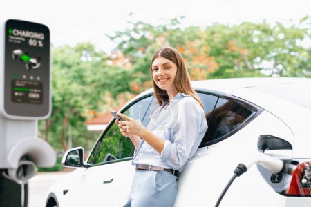 Photo for Young woman using smartphone online banking application to pay for electric car battery charging from EV charging station during vacation holiday road trip at national park or summer forest. Exalt - Royalty Free Image