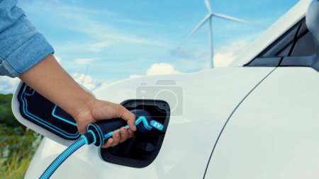 Téléchargez les photos : Insérer à la main le chargeur EV et recharger la voiture électrique de la station de charge avec un arrière-plan d'éolienne. Smart voiture écologique EV. Progrès technologique des énergies alternatives. Pertinence - en image libre de droit