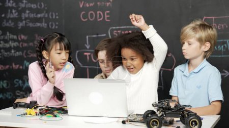 Photo for Multicultural smart children using laptop programing engineering code and writing or coding program in STEM technology classroom at blackboard written prompt. African girl raised hand. Erudition. - Royalty Free Image
