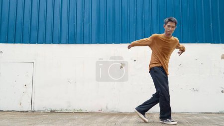 Photo for Hispanic man stretch arms and dance street dancing in front of wall. Motion shot of stylish dancer or choreographer in casual outfit practicing dancing in hip hop style. Outdoor sport 2024. Endeavor. - Royalty Free Image