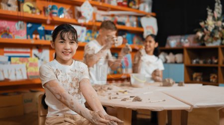 Photo for Cute girl smiling and sitting while diverse friend doing pottery workshop. Highschool student wearing dirty shirt while looking at camera at workshop in art lesson. Blurring background. Edification. - Royalty Free Image