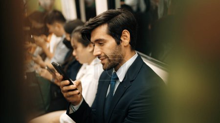 Téléchargez les photos : Homme d'affaires caucasien souriant regardant le téléphone portable ou jouant les médias sociaux tout en étant assis dans le train. Gestionnaire de projet attrayant se rendant sur le lieu de travail en utilisant les transports publics aux heures de pointe. Exultant. - en image libre de droit