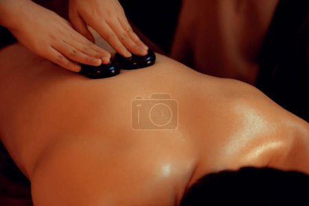 Photo for Hot stone massage at spa salon in luxury resort with warm candle light, blissful man customer enjoying spa basalt stone massage glide over body with soothing warmth. Quiescent - Royalty Free Image