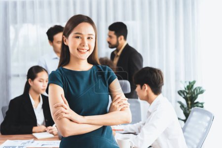 Téléchargez les photos : Jeune femme d'affaires asiatique pose avec confiance avec divers collègues dans le contexte occupé de la salle de réunion. Une équipe multiculturelle travaille ensemble à la réussite de l'entreprise. Portrait de dame de bureau. Concord - en image libre de droit