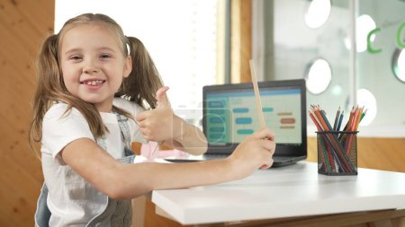 Photo for Smiling girl using laptop while look and show thumb up to camera. Cute child wearing headphone while working by using laptop writing code in STEM technology education. Online learning. Erudition. - Royalty Free Image