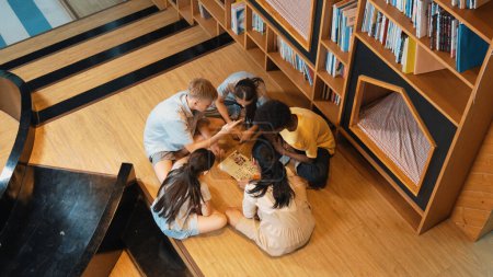 Photo for Top view of diverse children sitting in circle while looking at camera and waving hands at library surrounded with the stack of book. Multicultural highschool student greeting camera. Edification. - Royalty Free Image