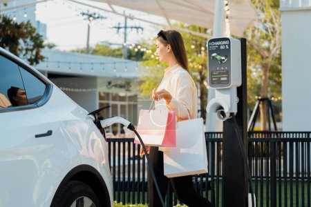 Photo for Young woman holding shopping bag recharge EV car battery from charging station at parking lot. Modern woman go shopping by environmental friendly electric vehicle in urban travel lifestyle. Expedient - Royalty Free Image