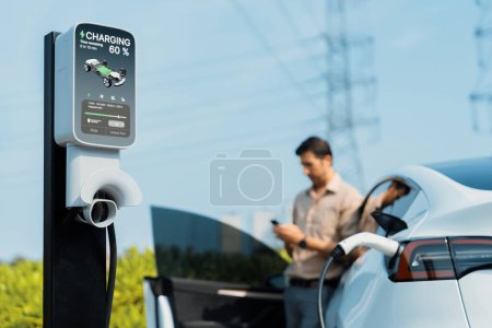 Photo for Man pay for electricity with smartphone while recharge EV car battery at charging station connected to power grid tower electrical as electrical industry for eco friendly car utilization.Expedient - Royalty Free Image