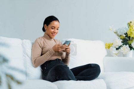 Téléchargez les photos : Jeune femme afro-américaine détendue utilisant un téléphone portable crucial sur le canapé dans le salon à la maison - en image libre de droit