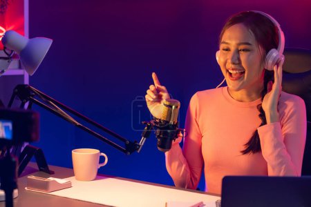 Photo for Host channel of young beautiful Asian broadcaster listening the music on social media with listeners, wearing pastel headphones, using microphone for singing at neon light indoor studio. Stratagem. - Royalty Free Image