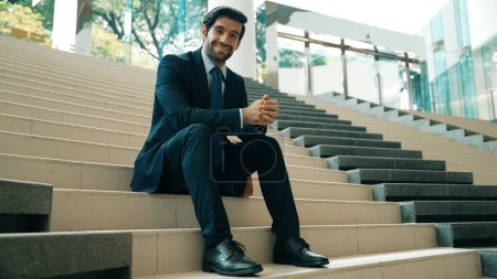 Photo for Smiling skilled businessman looking at camera while sitting on stairs. Young professional project manager smile at camera while holding mobile phone at outdoor with blurred background. Exultant. - Royalty Free Image