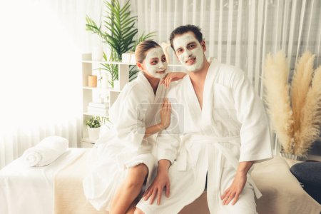 Téléchargez les photos : Couple heureux en peignoir avec masque crème visage profitant d'une ambiance sereine de spa salon resort ou hôtel pendant les vacances. Soin du visage choyante spa et soins de la peau avec relaxation essence. Tranquillité - en image libre de droit