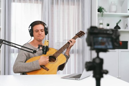 Photo for Host channel in smart singer recording by camera, playing guitar along singing, broadcasting on social media channel, wearing headphones to record video streamer at white modern studio. Pecuniary. - Royalty Free Image