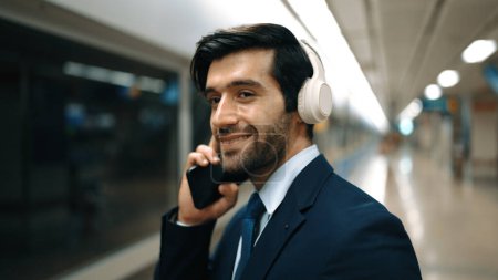 Photo for Smart business man calling phone while wearing headphone at train station. Professional executive manager talking to colleague about marketing strategy while waiting for train or subway. Exultant. - Royalty Free Image