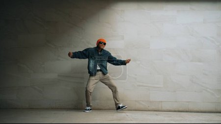 Foto de Motion shot of hipster realizar break dance en el fondo gris. Bailarina callejera practica el movimiento hip hop mientras usa un elegante paño de hip hop y se mueve a la música funky. Deporte al aire libre 2024. Rígidamente.. - Imagen libre de derechos