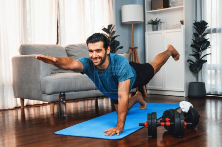 Téléchargez les photos : Homme flexible et dextérité en tenue de sport faisant position de yoga en posture de méditation sur tapis d'exercice à la maison. Mode de vie sain de yoga de gaieté à domicile avec l'esprit paisible et la sérénité. - en image libre de droit