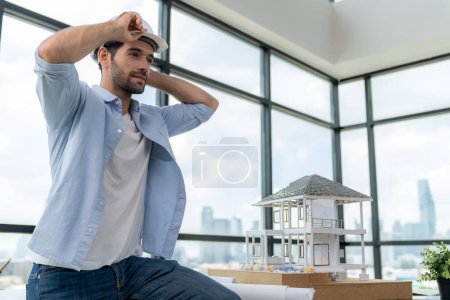 Photo for Portrait of skilled architect engineer wear safety helmet while sitting on table with house model, project plan and architectural equipment surrounded by skyscraper view. Civil engineering. Tracery. - Royalty Free Image