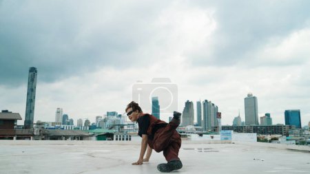 Téléchargez les photos : Tournage en mouvement de la performance de danse B-boy par un danseur de rue professionnel sur le toit avec gratte-ciel, vue sur la ville. Attrayant hipster asiatique montrer pas énergique. Sport de plein air 2024. Endeavor. - en image libre de droit