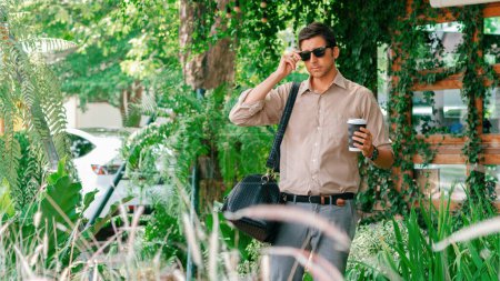 Foto de Joven hombre guapo salir de la cafetería con cafetería jardín, sosteniendo una taza de café y una bolsa de compras. Moderno feliz despreocupado con gafas de sol estilo de vida. Expediente - Imagen libre de derechos