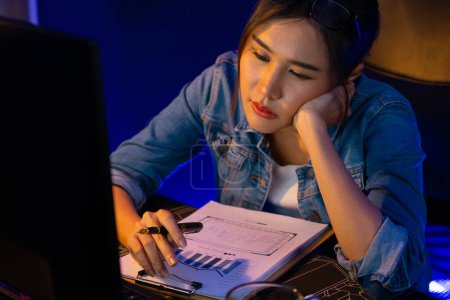 Photo for Exhausted young beautiful Asian creative feeling sleepy, looking on pc to wait email sending back with comparing data sheet of project creator social media online at neon office at night. Stratagem. - Royalty Free Image