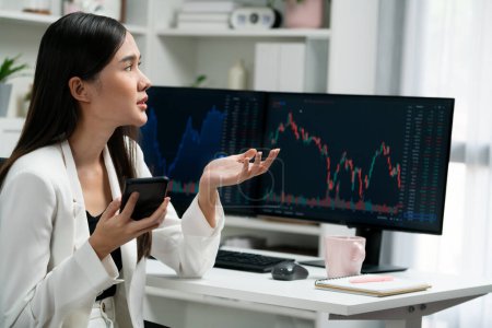 Stressful young beautiful Asian businesswoman focusing on smartphone, analyzing possible risk to profit or loss in dynamic exchange stock market graph on pc screen at modern office. Stratagem.