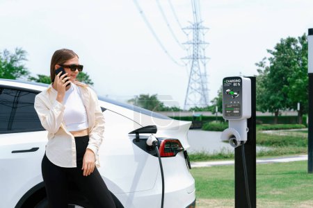 Photo for Young woman recharging EV car battery while talk on phone at charging station connected to electrical power grid tower facility as electrical industry for eco friendly vehicle utilization. Expedient - Royalty Free Image