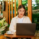 Young woman working on laptop at outdoor cafe garden during springtime, enjoying serenity ambient at coffee shop. Digital nomad freelancer or college student working remotely or blogging. Expedient