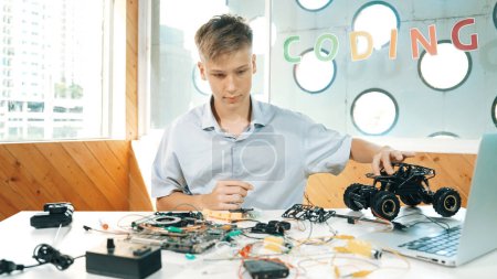 Photo for Highschool boy fixing car model with laptop and electric tool placed on table. Closeup of teenager inspect robotic machine construction while using eletronic equipment in STEM classroom. Edification. - Royalty Free Image
