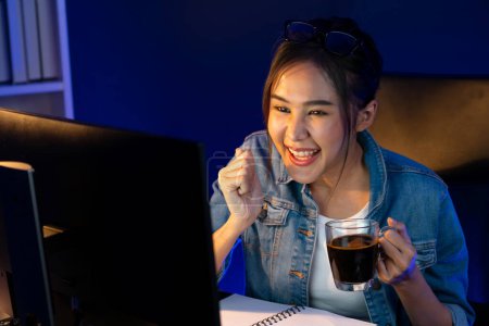 Photo for Smiling young beautiful Asian creative officer looking on pc in sales target online while holding coffee cup at neon light room at night time, earning jobs freelancer strategy marketing. Stratagem. - Royalty Free Image