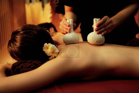 Photo for Hot herbal ball spa massage body treatment, masseur gently compresses herb bag on couple customer body. Serenity of aromatherapy recreation in warm lighting of candles at spa salon. Quiescent - Royalty Free Image