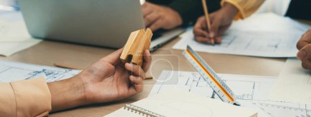 Photo for Professional architect team discussion about architectural project on meeting table with blueprint and wooden block scatter around at modern office. Closeup. Focus on hand. Delineation. - Royalty Free Image