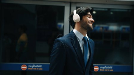 Photo for Closeup of smart executive manager listen relaxing music while waiting at train station. Happy male leader wear headphone and standing at door area. Public transport concept. Dark filter. Exultant. - Royalty Free Image