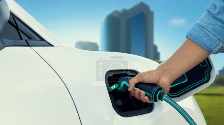 Téléchargez les photos : Insérer à la main le chargeur EV et recharger la voiture électrique de la station de charge affichant un hologramme futuriste de l'état de la batterie sur fond de parc urbain vert ESG. Énergie propre durable intelligente. Pertinence - en image libre de droit