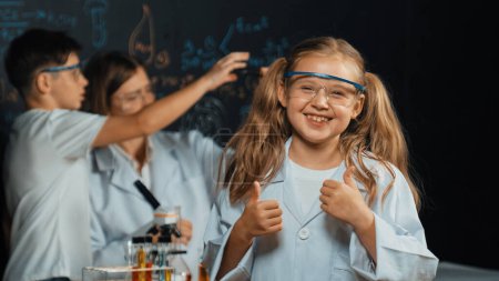 Photo for Smart caucasian girl showing thumb while teacher and student doing experiment behind. Child looking at camera while boy looking under microscope at black board written chemical theory. Erudition. - Royalty Free Image