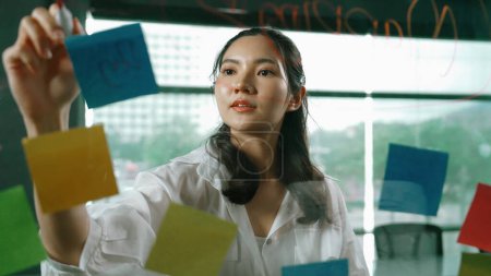 Photo for Professional businesswoman writing marketing idea on glass board. Smiling young beautiful project manager looking at colorful sticky notes with show creativity marketing strategy. Manipulator. - Royalty Free Image