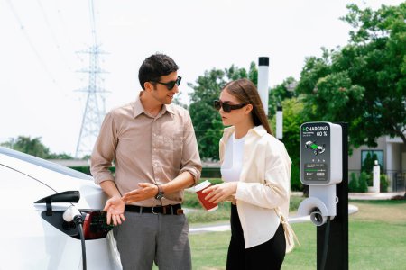 Photo for Young couple with coffee pay for electricity while recharge EV car battery at charging station connected to electrical power grid tower as electric industrial for eco friendly car travel. Expedient - Royalty Free Image