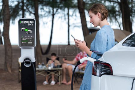 Photo for Outdoor adventure and family vacation camping at sea travel by eco friendly car. Woman or mother check cars battery with smartphone while charging EV car frin charging station in campsite. Perpetual - Royalty Free Image