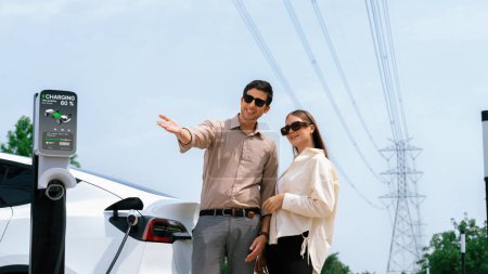 Téléchargez les photos : Jeune couple rechargent la batterie de voiture EV à la station de charge connectée à l'installation industrielle électrique de tour de réseau électrique comme industrie électrique pour l'utilisation de véhicule respectueuse de l'environnement. Expedient - en image libre de droit