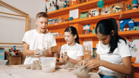 Photo for Happy diverse highschool student modeling clay at pottery workshop. Cute children with mixed raced working and making cup of clay by using carving equipment at art lesson. Creative class. Edification. - Royalty Free Image