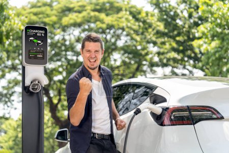 Photo for Man recharging battery for electric car during road trip travel EV car in natural forest or national park. Eco friendly travel during vacation and holiday. Exalt - Royalty Free Image